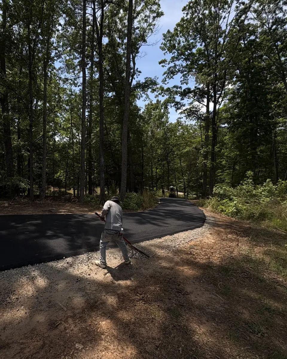 Crew member hand-raking the edge of a new asphalt driveway through the trees